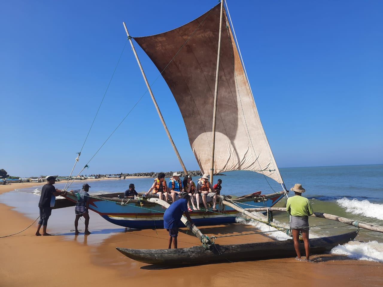 Tourists in life vests standing on beach beside traditional oruwa with large triangular sail against clear blue sky
